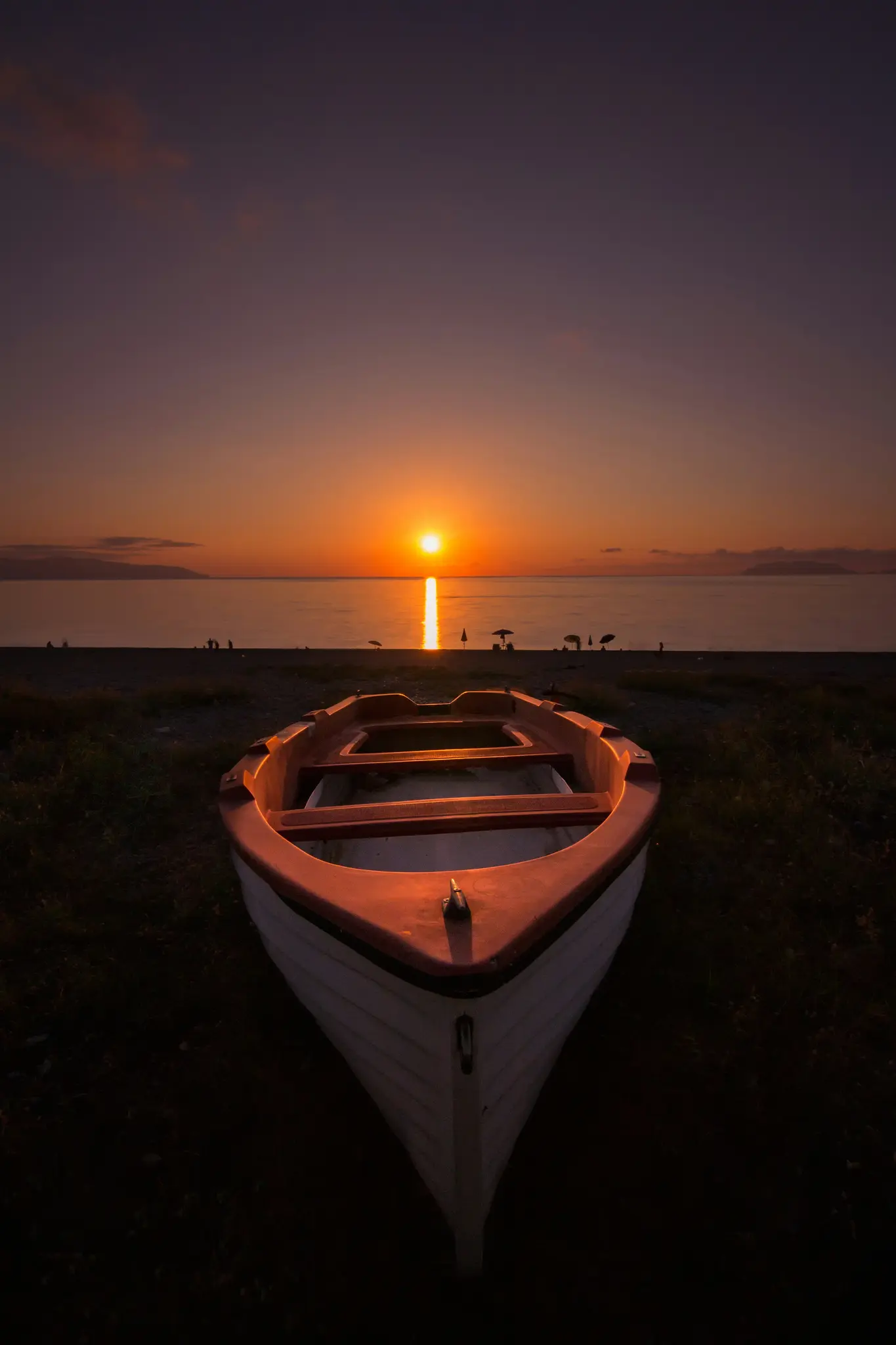 Un pequeño barco blanco y naranja descansa en una playa oscura, de cara al mar. El sol se pone justo delante, proyectando un