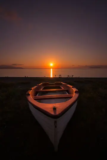 En lille hvid og orange båd hviler på en mørk strand, vendt mod havet. Solen går ned lige forude og kaster en orange glød.