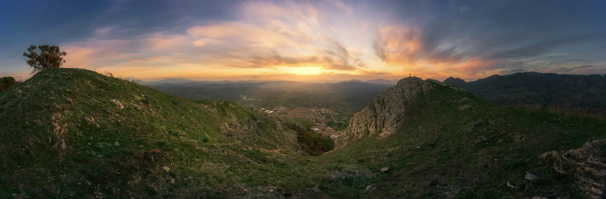 Et bredt panoramabillede af et bjerglandskab ved solnedgang. Himlen er fyldt med orange, gule og blå skyer.