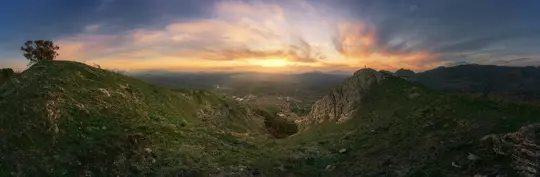 Et bredt panoramabillede af et bjerglandskab ved solnedgang. Himlen er fyldt med orange, gule og blå skyer.