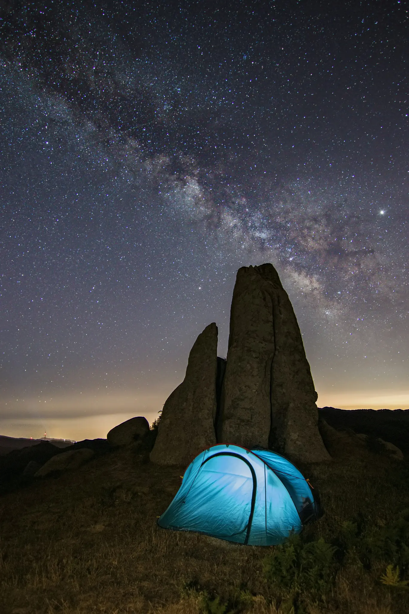 Una tenda blu vibrante brilla dall'interno, posizionata in un paesaggio roccioso sotto un cielo notturno limpido pieno di Via
