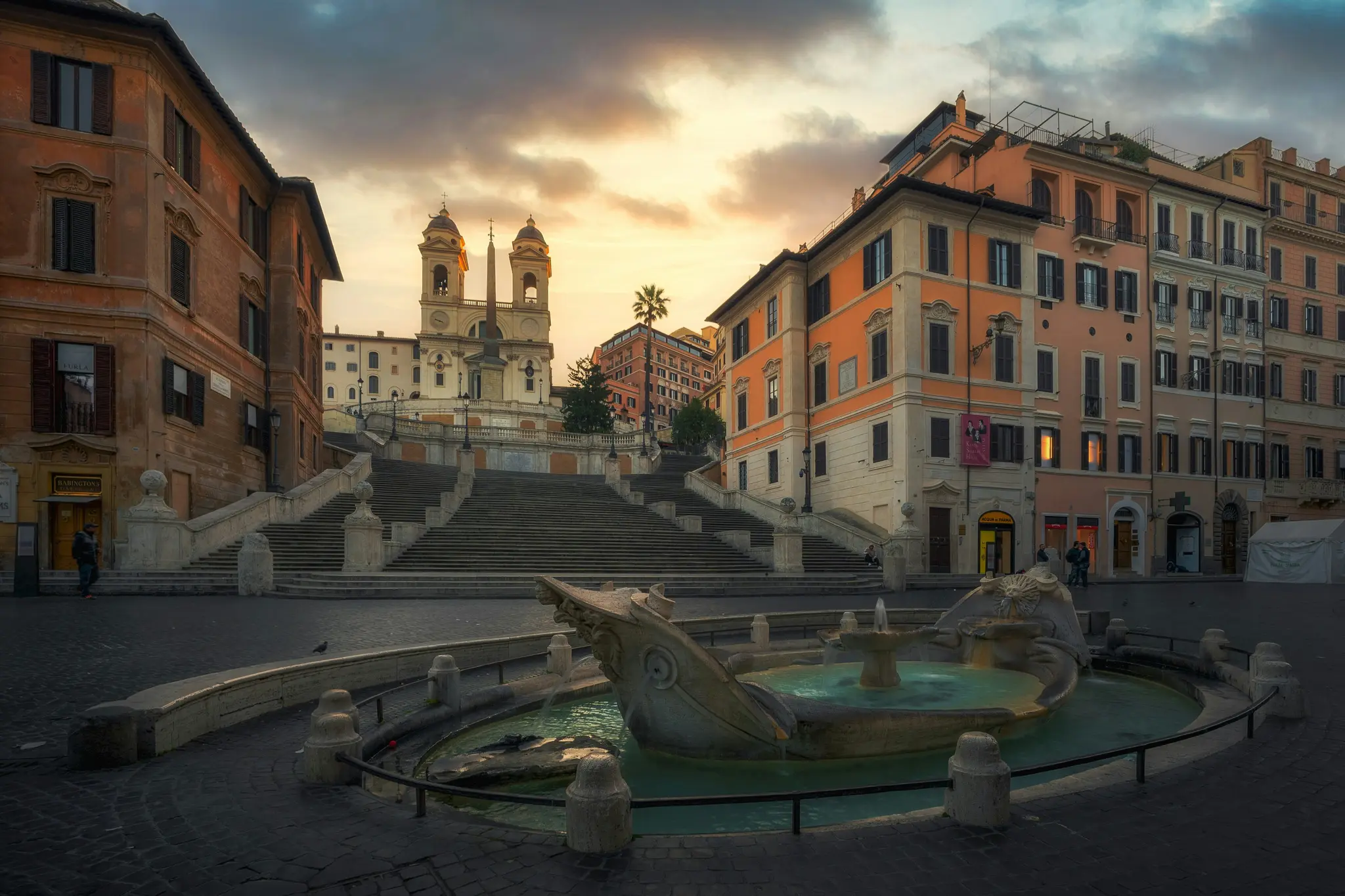 En vidvinkelbild av Spanska trappan och Fontana della Barcaccia i Rom vid solnedgång. Himlen är varm med orange och gula