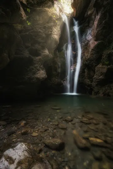 Et langtidseksponeringsfoto af et vandfald, der styrter ned i en klar, lav pool omgivet af mørke, teksturerede klippevægge.