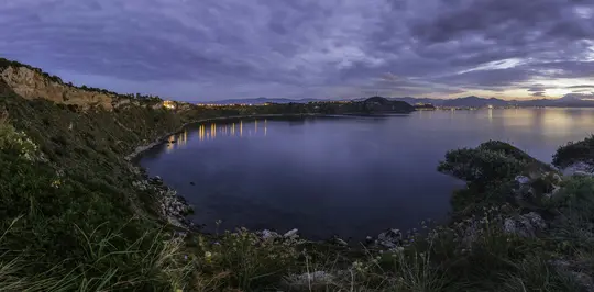 Et panoramisk landskabsfoto ved skumringstid viser en rolig bugt med mørkeblåt vand, der reflekterer orange bylys fra den