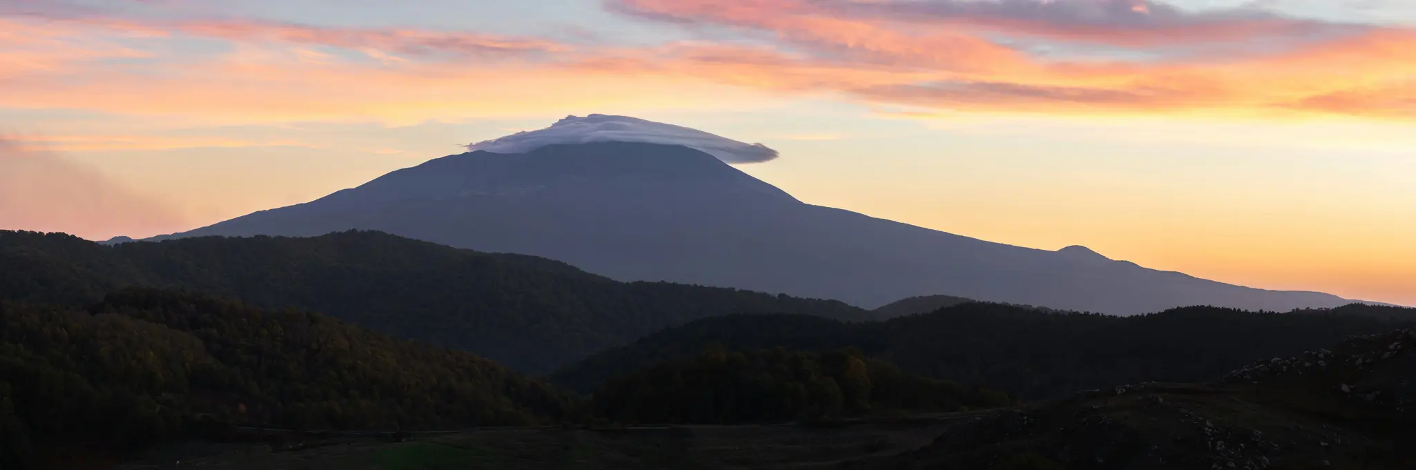 Široký panoramatický záběr velké, tmavé hory s mrakem na vrcholu, na pozadí zářivé oranžové a růžové oblohy při západu