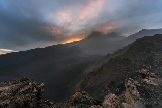 Et bredt landskabsbillede af mørke, barske vulkanske bjerge under en dramatisk solnedgangshimmel. Orange og lyserødt lys