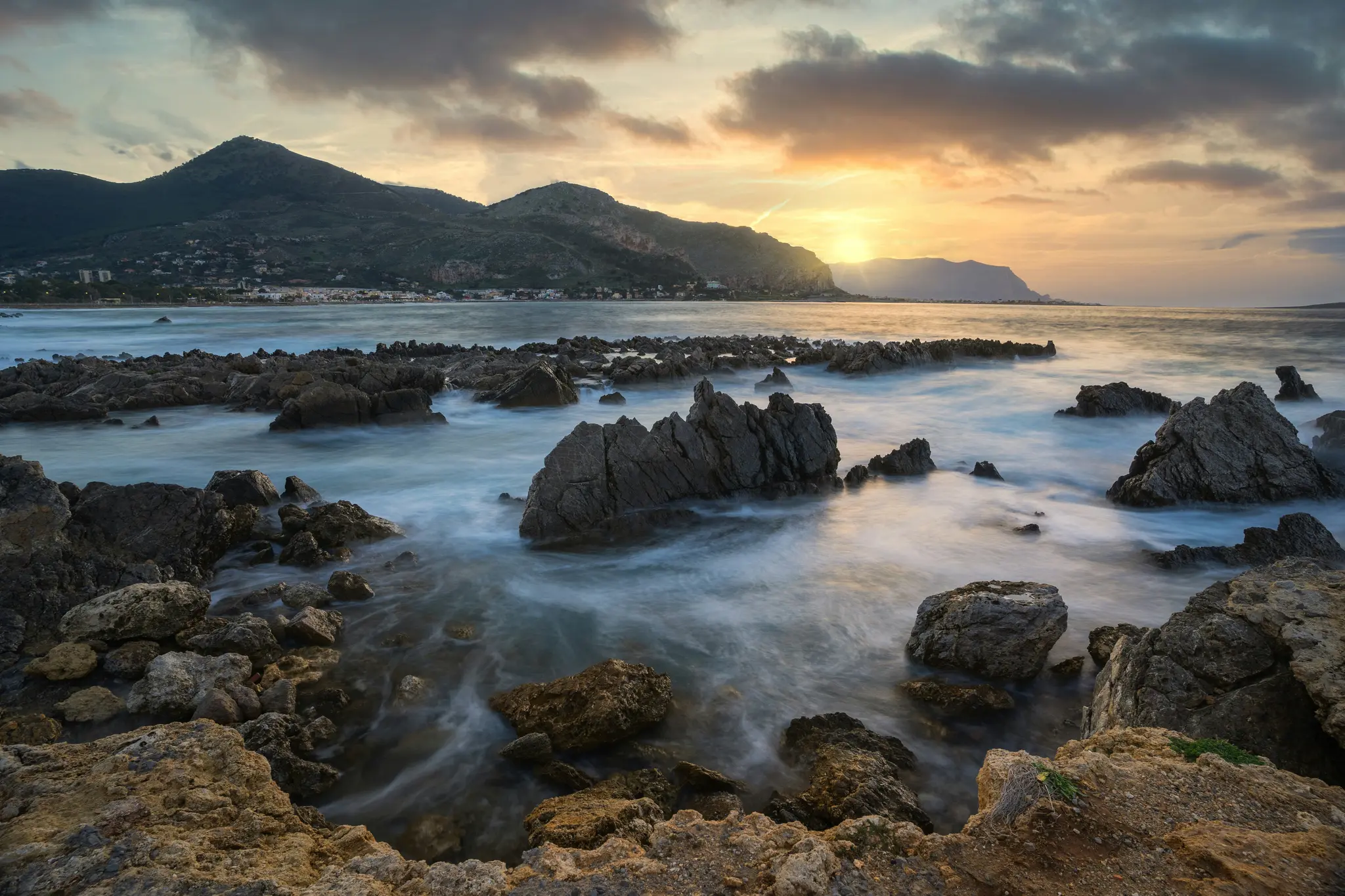 Un'ampia foto panoramica di una costa rocciosa al tramonto. Il sole tramonta dietro le montagne in lontananza, proiettando un