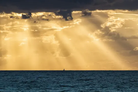 En lille sejlbåd er silhuet på horisonten af et dybblåt hav, under en dramatisk himmel med gyldne solstråler.