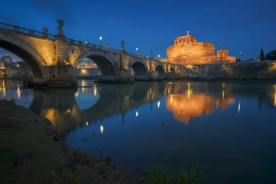 En natudsigt over det oplyste Castel Sant'Angelo og Ponte Sant'Angelo med deres refleksioner synlige i Tiberfloden.