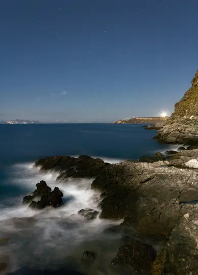 Et langtidseksponeringsbillede af en mørk, klippefyldt kyst under en stjerneklar nattehimmel. Havet er dybblåt, og bølger