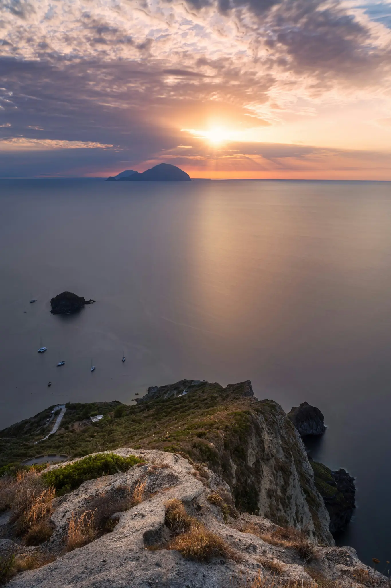 Krajinářská fotografie z ptačí perspektivy zachycující západ slunce nad klidným mořem. Slunce, jasně oranžová koule, je nízko