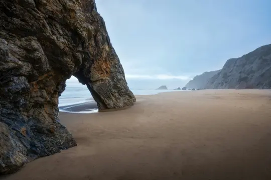 En stor, mørk klippebue står på en bred, sandet strand. Bølger bryder gennem buen, og klipper afgrænser den fjerne kyst.