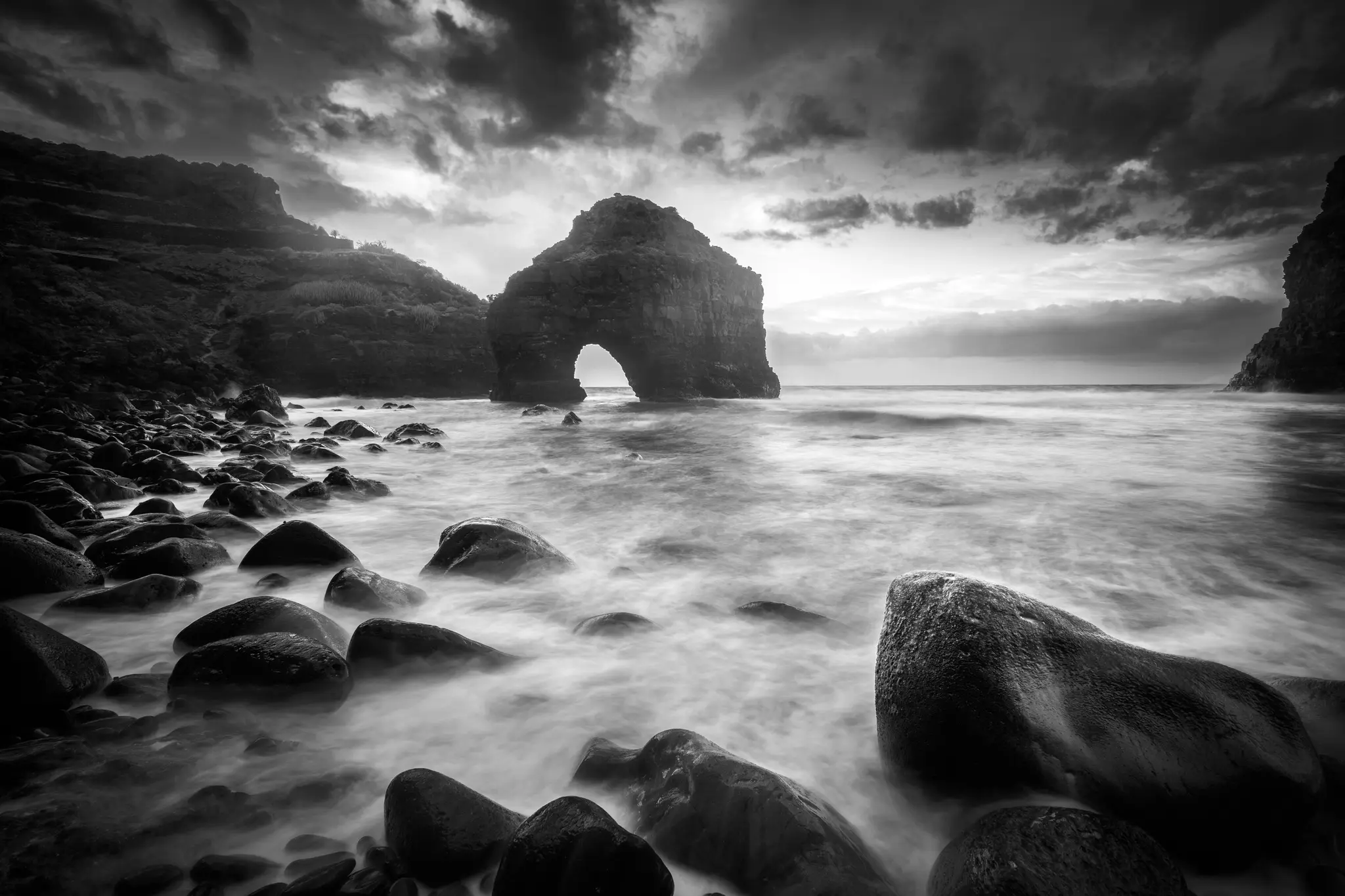 Una drammatica esposizione lunga in bianco e nero di una costa rocciosa. Un grande arco di roccia naturale si trova