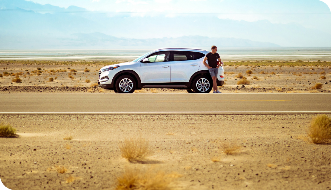 A driver leaning on a car parked on a desert road, surrounded by open, sandy scenery