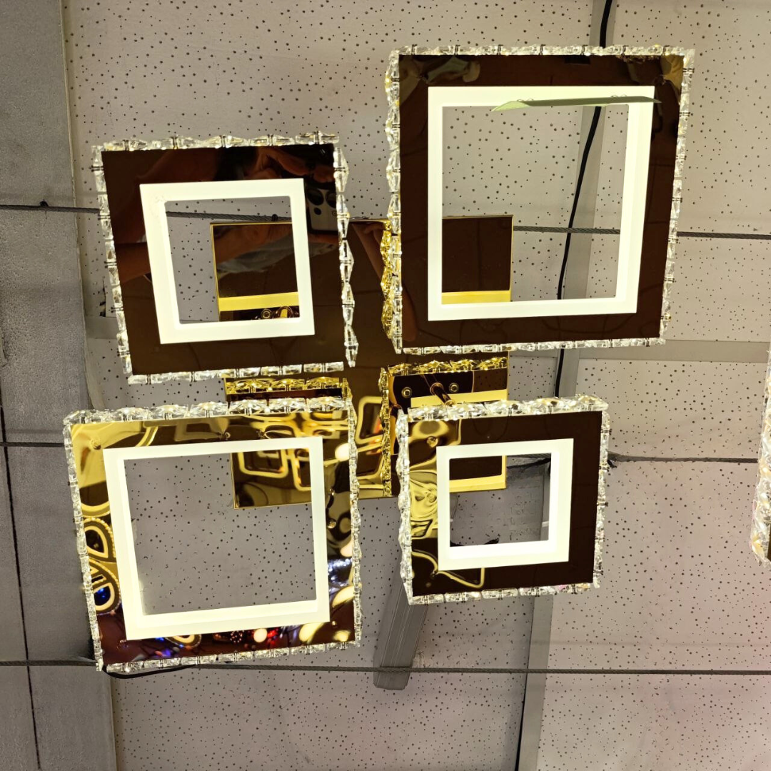 Сучасна квадратна світлодіодна люстра з прозорим акрилом і золотим корпусом, чотири світлових сегменти з кришталевими гранями, фото з виставки освітлення, актуальний дизайн для вітальні або кухні, преміальний світильник у стилі модерн, крупний план під стелею, інтер’єр магазину, відблиски на глянцевих поверхнях