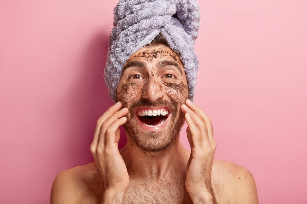Male beauty concept. Happy joyous man applies coffee scrub on face, removes dark dotes, wants to look refreshed, has wrapped towel on head, poses against pink background topless, shows white teeth