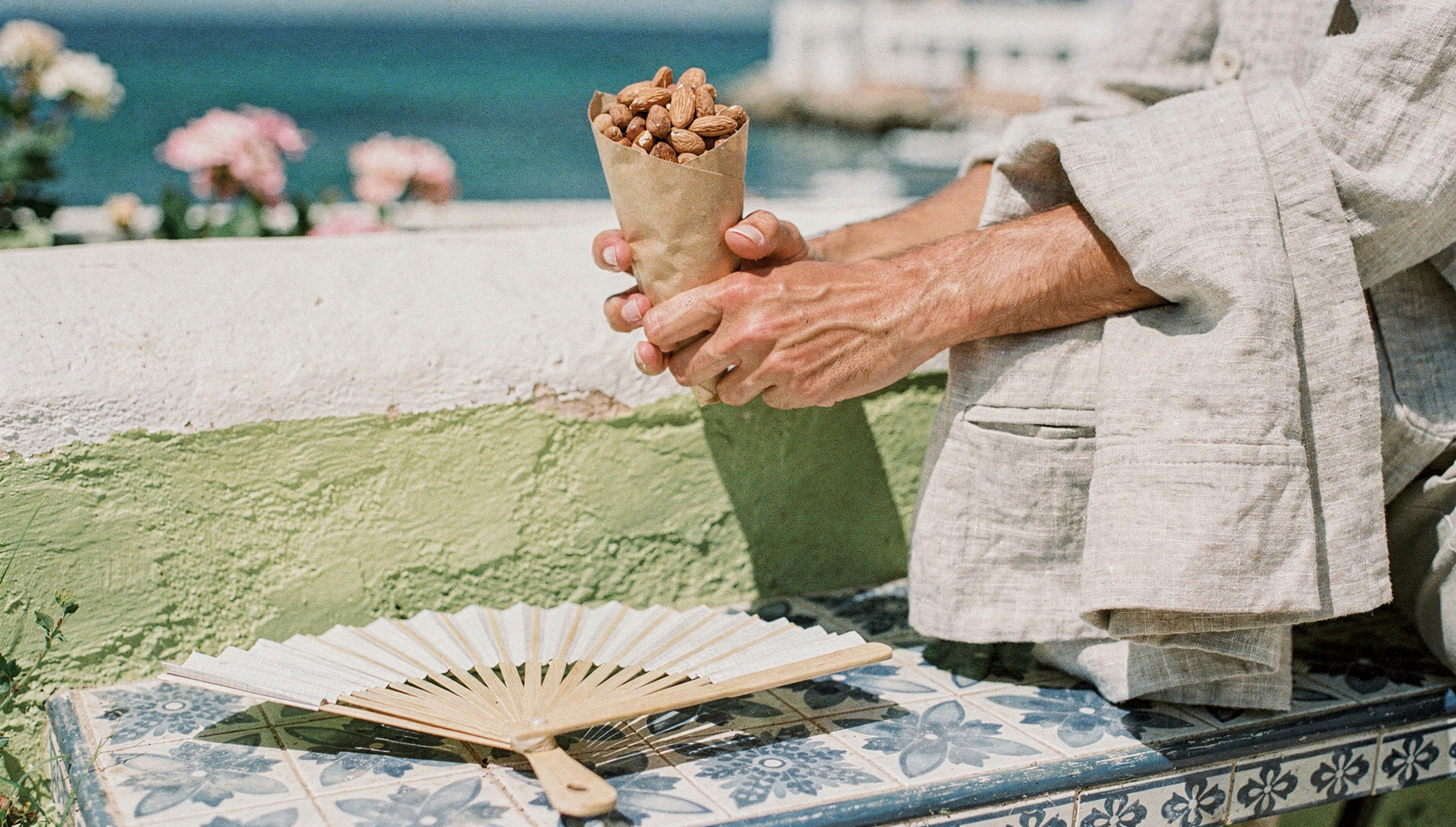 A hand holding a paper cone of roasted almonds beside a tiled bench