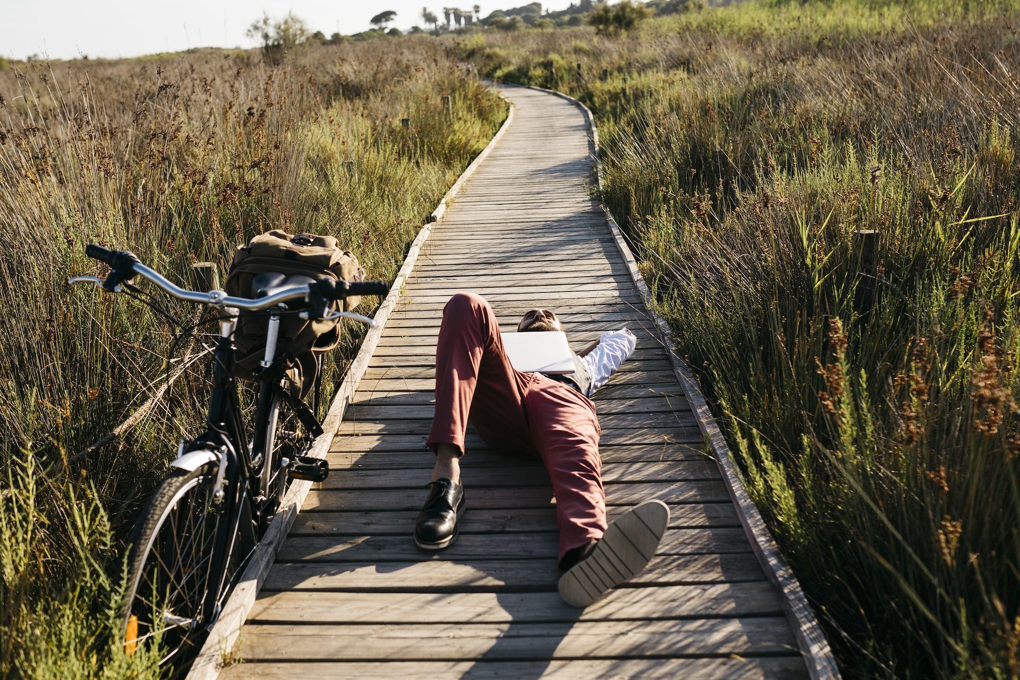 Well dressed man with laptop lying on a wooden walkway in the countryside next to a bike