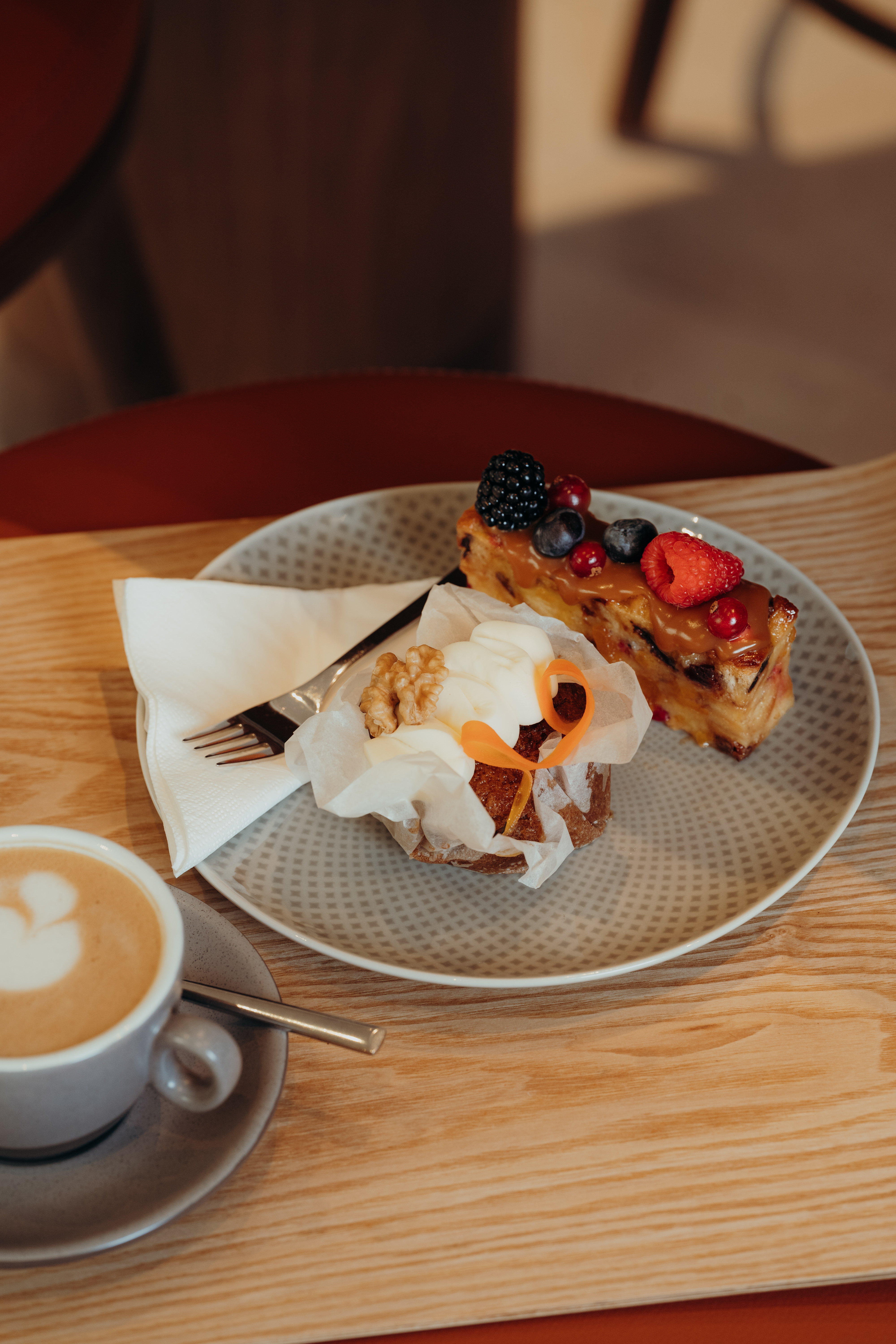 Ein Teller mit einem Stück Kuchen, garniert mit frischen Beeren, und einem Muffin mit Sahne und einer Walnuss, daneben eine Serviette und eine Gabel. Neben dem Teller steht eine Tasse Cappuccino mit Latte Art auf einem Holztisch.