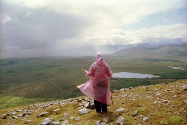 Photograph of a woman in a pink rain coat looking at the view from a hill/mountain with a lake in the distance