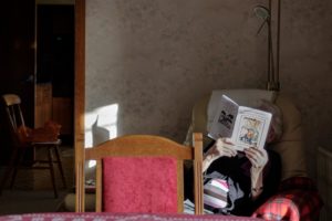 Older woman sitting in a chair with a book covering her face in the corner of living room.