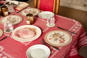 Table set with a red table cloth and placemats. On the middle of the table is a plate of sliced ham and a jar of chutney waiting to be eaten.