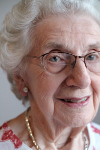 Close up portrait of an older woman looking directly to the camera. She has glasses, a small gold necklace and pearl earrings on.