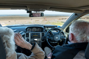 Taken from inside the back of a car looking out onto a beach. In the foreground is in the inside of the car with two people looking out; one older woman using binoculars to look out to the right, whilst an older man looks straight ahead and the reflection of his face has been caught in the rear view mirror.