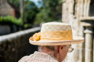 Back of a womans head wearing a straw hat and textured beige top in the middle of the frame. In the background are green trees and a light coloured stone building.