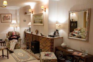 A couple of older people asleep on a sofa and arm chair in a living room. The pair are apart, with the man asleep in the chair whilst the reflection of the woman can be seen in a mirror. There is a fireplace and rug in the middle of the room along with various ornaments and paintings around the room.