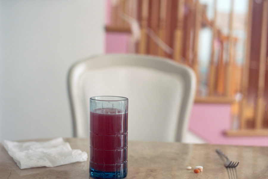 Photograph of a blue glass with raspberry iced tea, a napkin, vitamins, and a fork set at a marbled table with a chair and a set of stairs behind it.