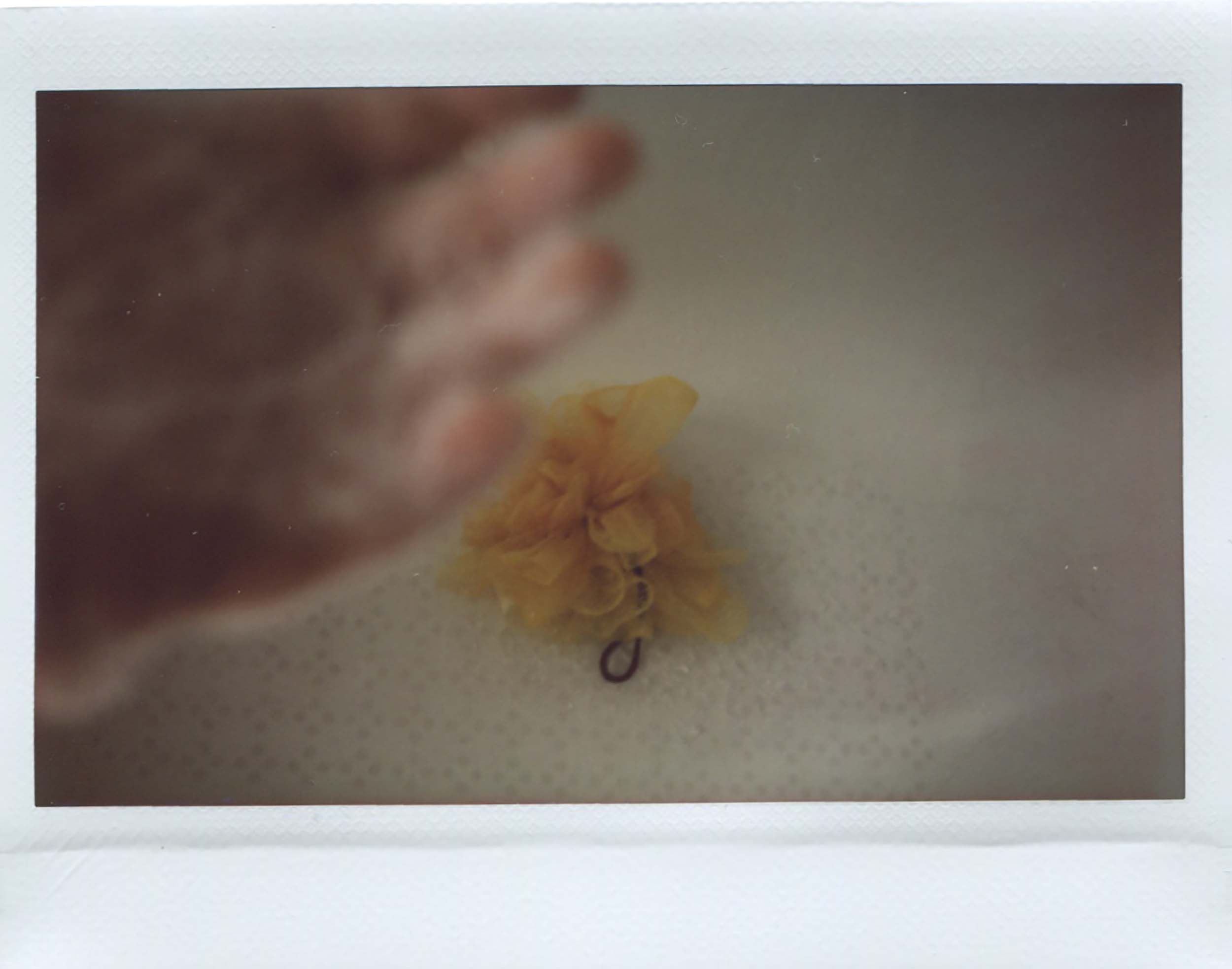 Instax Wide picture taken over a bath, left hand of the artist in the top left corner, out of focus but can see the bubbles. There's an orange body puff in the centre of the image.