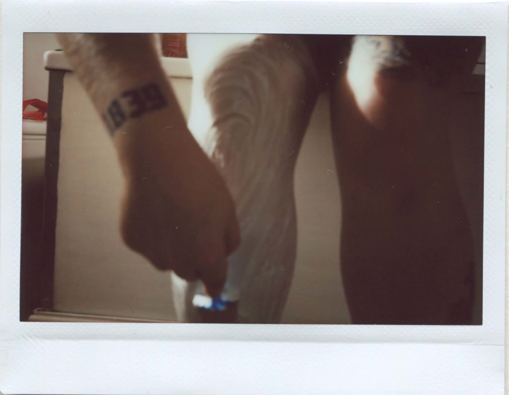Instax wide photo of the artists lower legs and right arm. The right leg is covered in shaving foam and she is applying a razor to it
