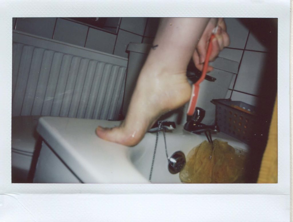 Instax wide print showing the artist's foot on the corner of the bath in the middle of the frame. She is holding a foot file to her heel.