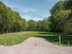 Photograph of a gravel path blocked off by a chain fence and a single traffic cone, with a field and trees behind and blue sky above