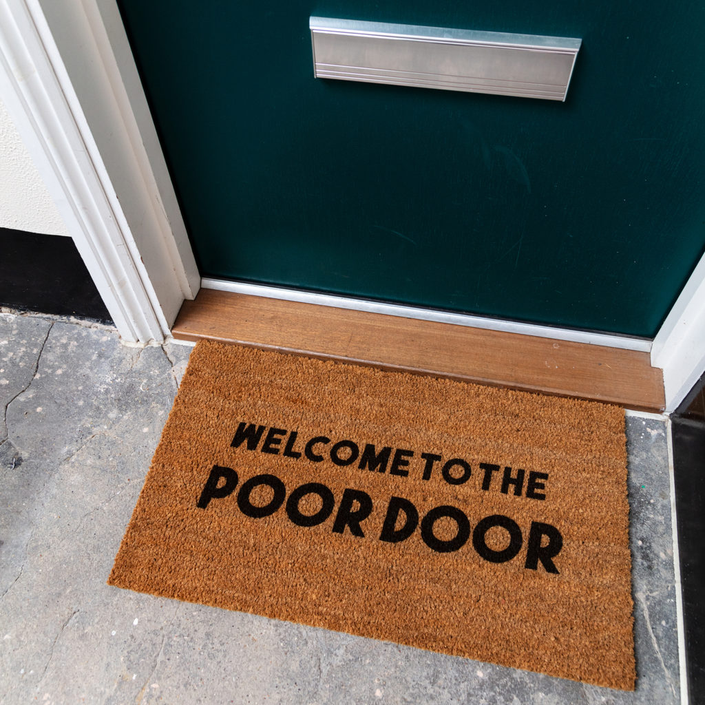 Photograph of a door mat outside a flat door with welcome to the poor door printed on it.