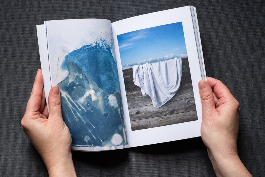Photograph of hands holding a book open on a dark grey background. On the left page is a cyanotype and on the right is a photograph of a white sheet draped on a wooden fence, with blue sky above.