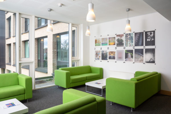 Photograph of a seating area with green sofas in Art at the ARB at University of Cambridge, with poster prints displayed on the wall and exterior windows visible through the large window to the left