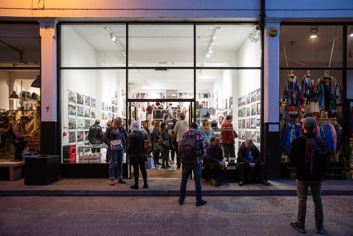 A picture containing an art gallery at night, road, walking people.