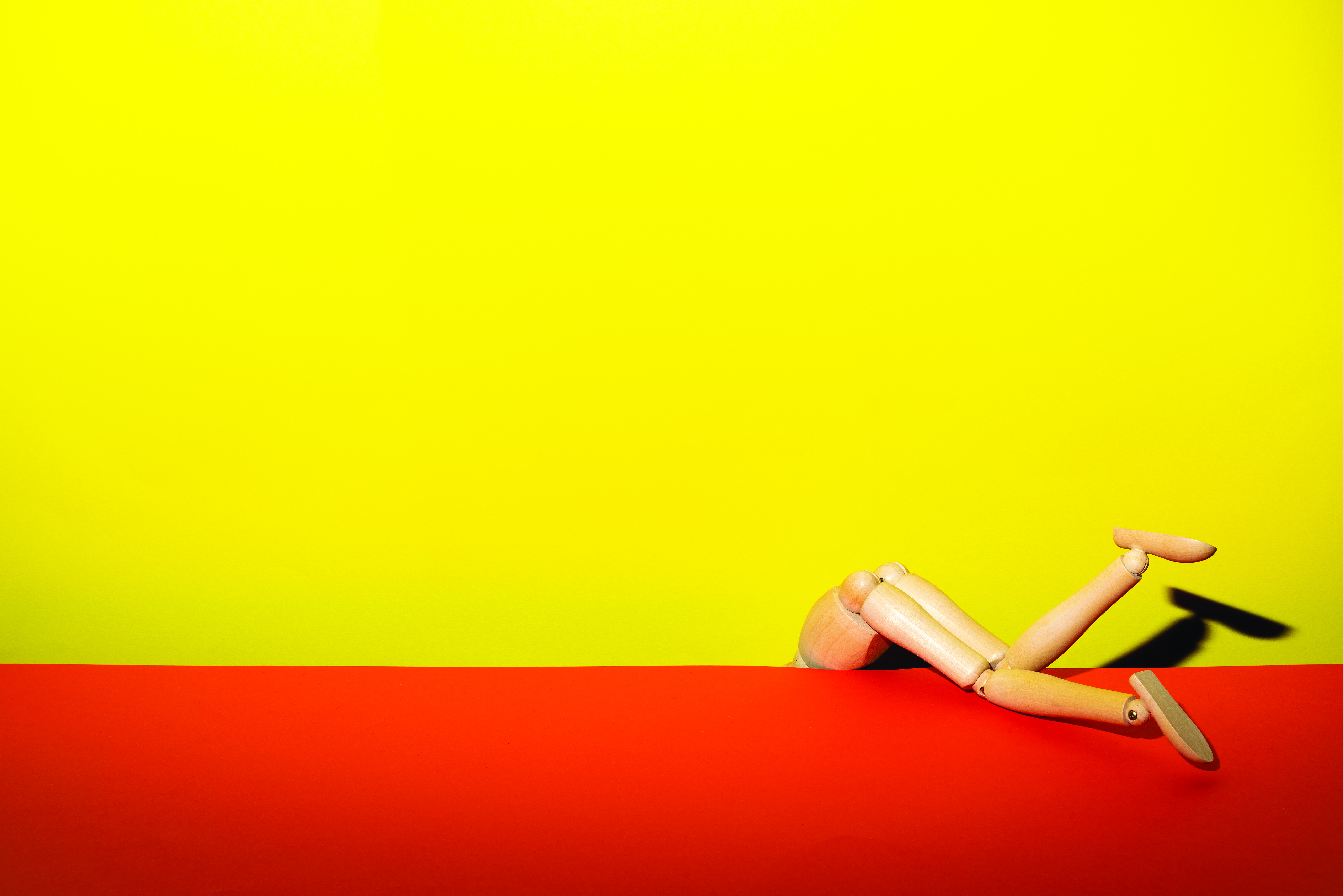 A wooden artist's model in the shape of a person, legs emerging from between a yellow wall and red floor, reenacting an image by Guy Bourdin