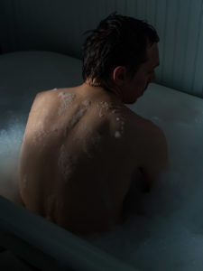 Photograph of a person sitting upright in a bubble bath with their back to the camera, mostly in darkness, with light hitting their left shoulder