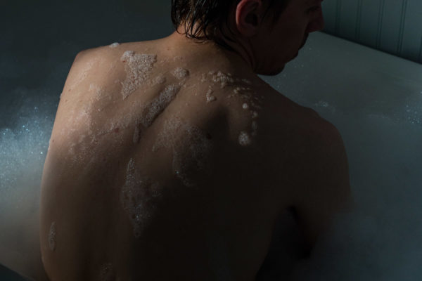 Photograph of a person sitting upright in a bubble bath with their back to the camera, mostly in darkness, with light hitting their left shoulder