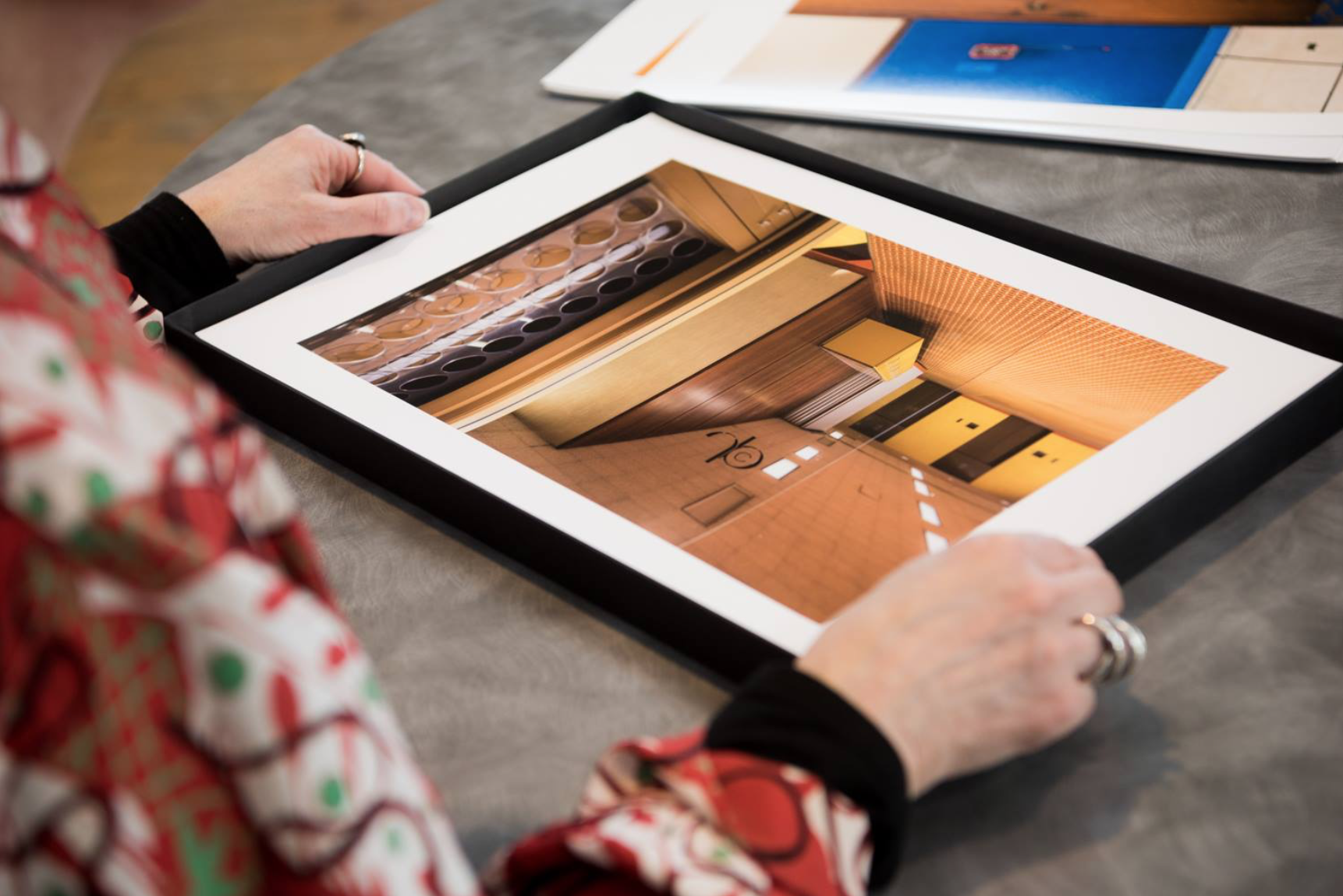 Photograph taken from behind and to the right of a person holding a framed print of a photograph of an interior on a table, during a portfolio review.
