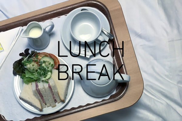 Photograph looking down on a tray with triangular ham sandwiches and a side salad, a small jug of milk, cup and saucer and small teapot, with the words 'LUNCH BREAK' in the centre