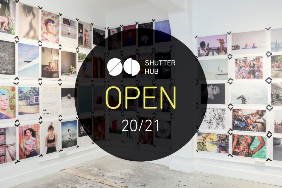 Graphic with a photograph of newspaper poster printed photographs displayed taped to the walls at London's Old Truman Brewery, with a black circle in the centre containing the Shutter Hub logo, with 'OPEN' and '20/21' below.