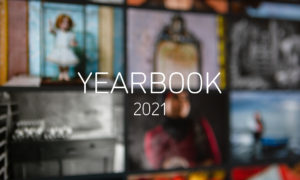 Graphic with an out of focus photograph of a computer monitor displaying the YEARBOOK 2020 online exhibition, with white text in the centre reading'YEARBOOK 2021'