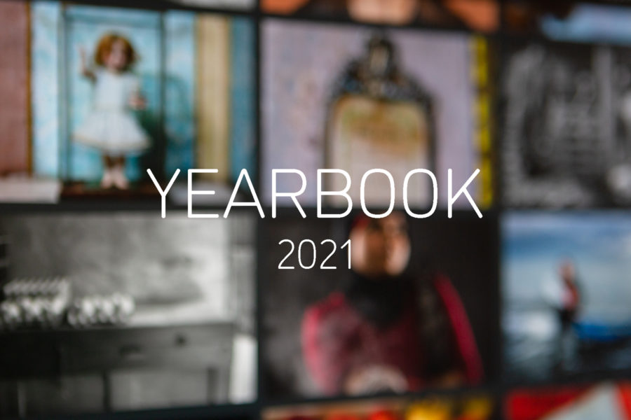 Graphic with an out of focus photograph of a computer monitor displaying the YEARBOOK 2020 online exhibition, with white text in the centre reading'YEARBOOK 2021'