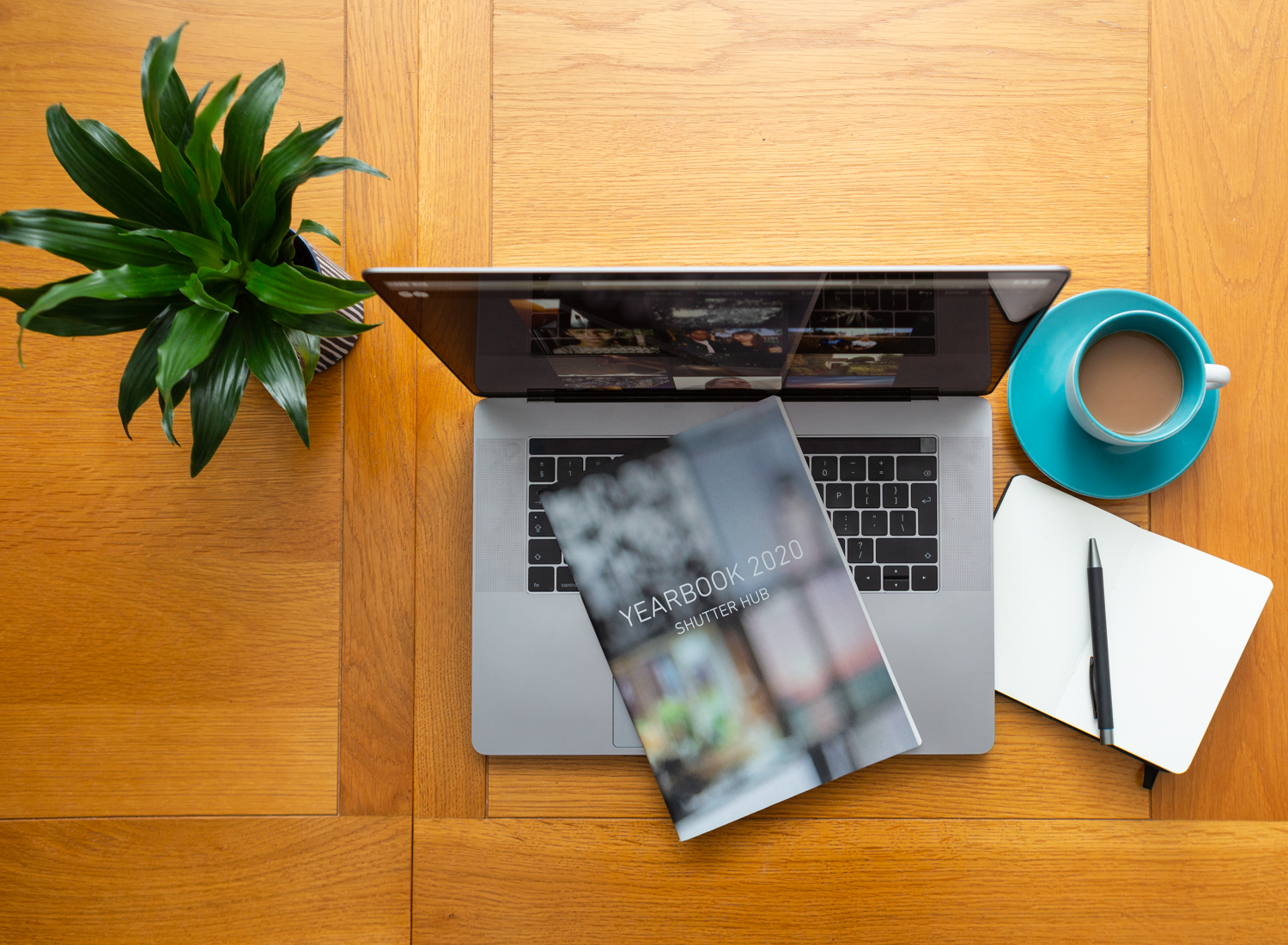 Looking down at a table with a pot plant to the left, laptop with YEARBOOK printed publication on top, a cup of coffee and a notebook with a pen to the right