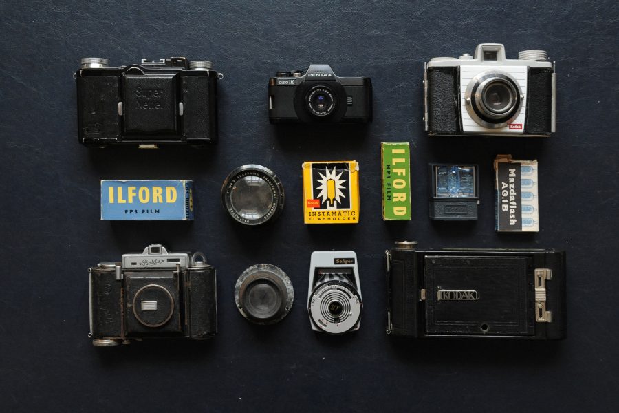 Photograph of vintage camera equipment arranged on a black background, including cameras, film and a light meter. The word ‘Ilford’ can be read on two boxes of film towards the centre of the frame.
