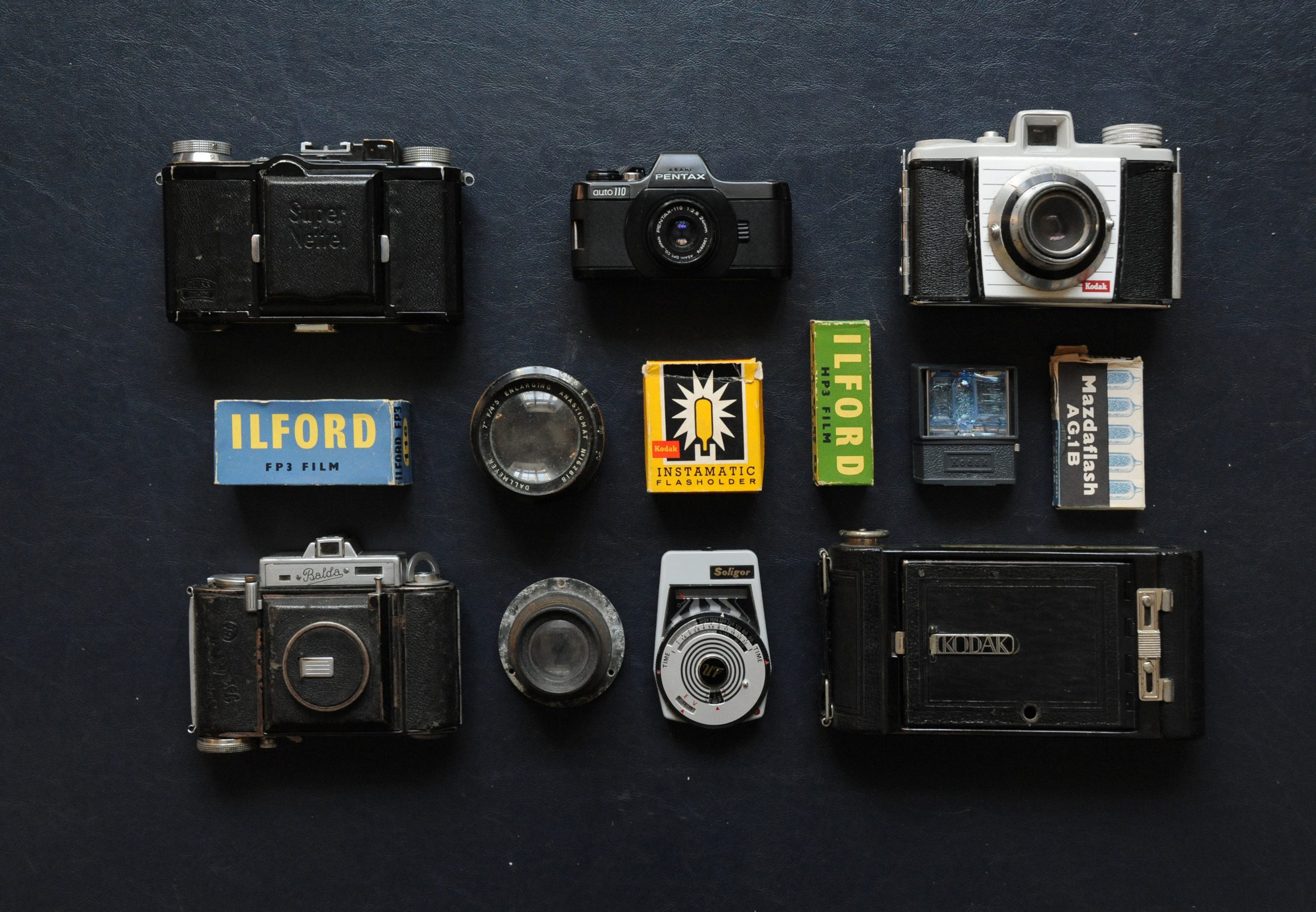 Photograph of vintage camera equipment arranged on a black background, including cameras, film and a light meter. The word ‘Ilford’ can be read on two boxes of film towards the centre of the frame.